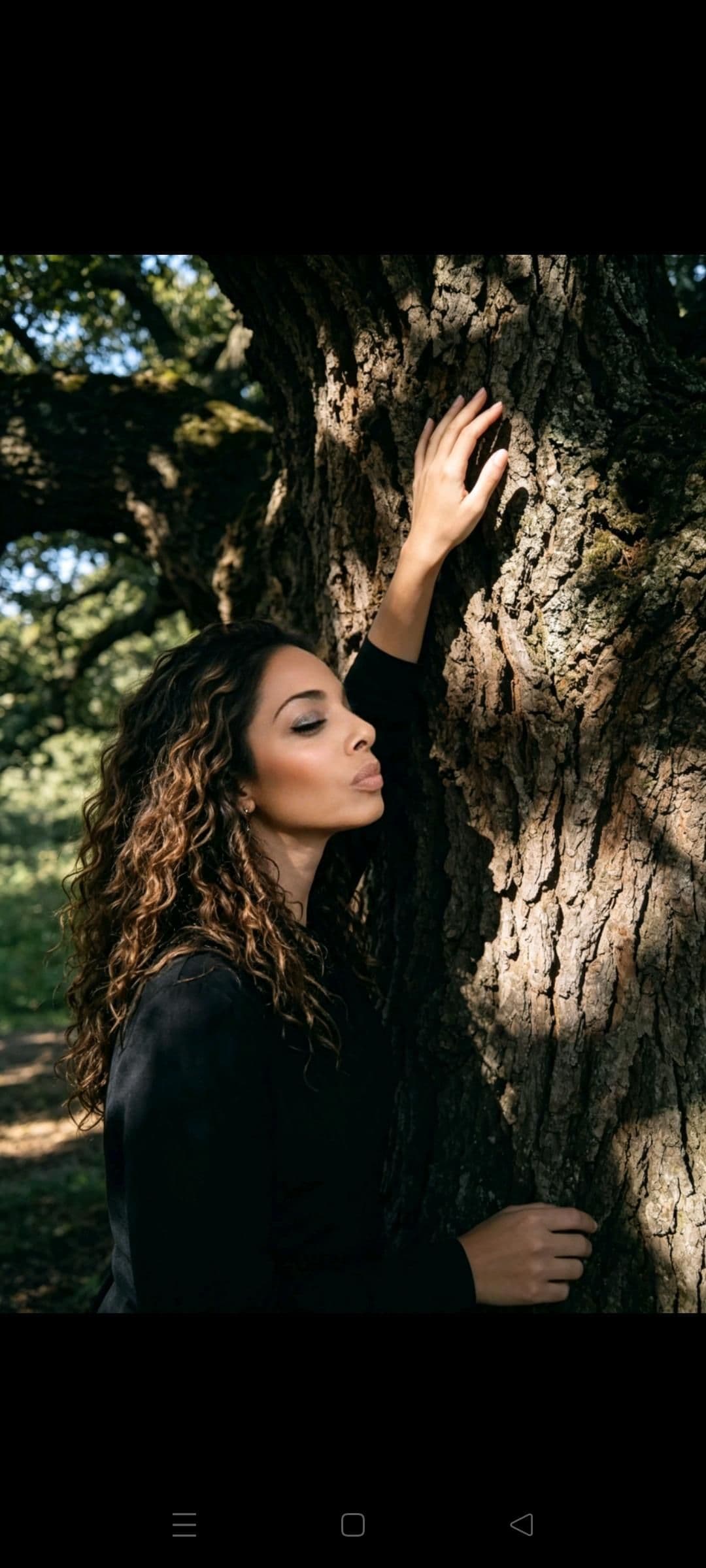 Woman with long curly hair leans peacefully against a large tree trunk in dappled sunlight.