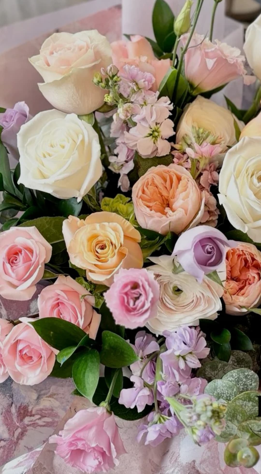 Pastel bouquet featuring white, pink, and peach roses, lavender stock, and fresh green foliage.